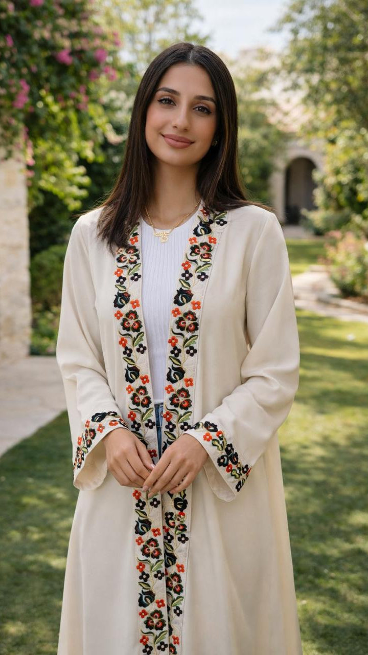 A woman stands outdoors in a garden wearing a long cream-colored embroidered jacket over a white top. The jacket features colorful floral embroidery in red, orange, black, and green along the front opening, cuffs, and a central hanging detail.