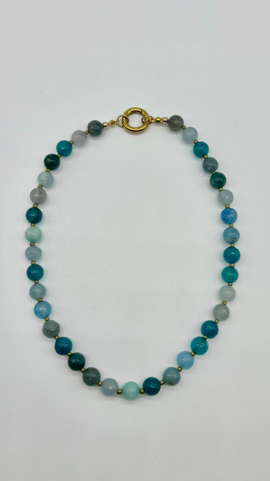 A beaded necklace made of round polished beads in shades of teal, blue, green, and gray, separated by small gold spacers and finished with a gold clasp, shown on a plain light background.