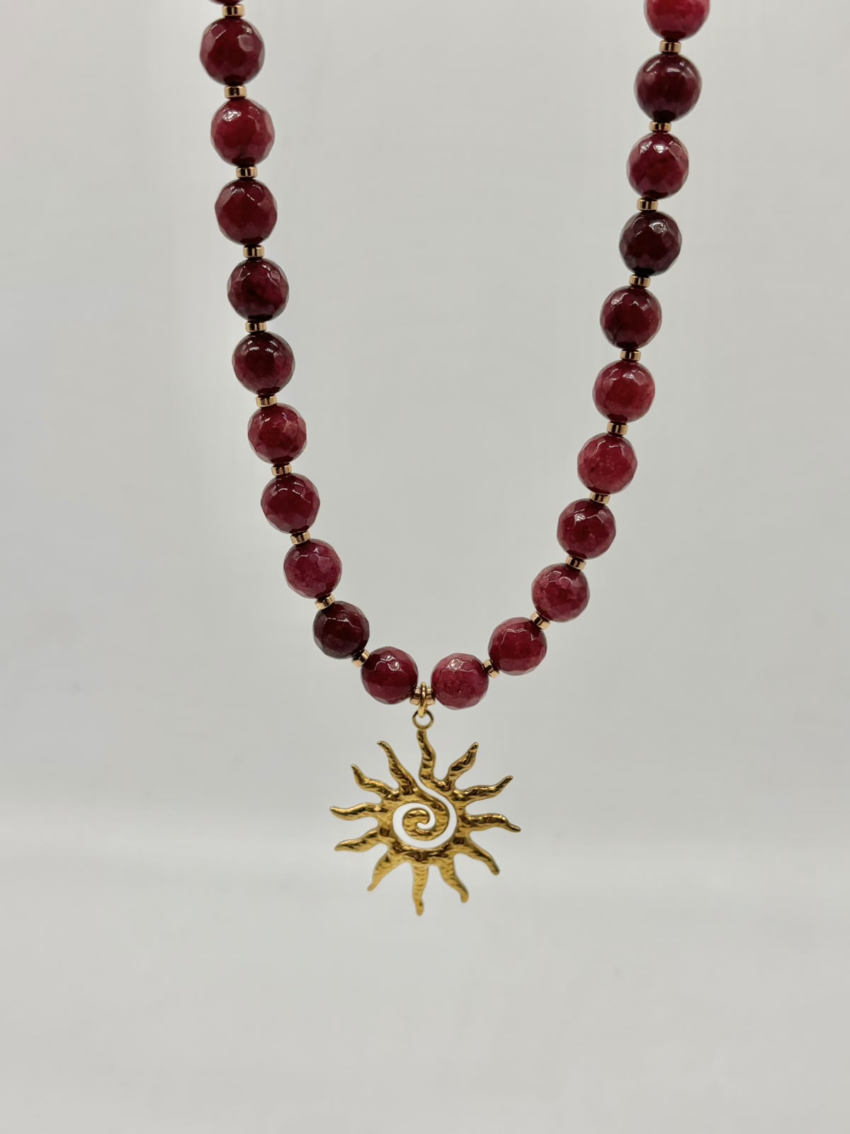 A beaded necklace featuring round, polished deep red beads separated by small gold spacer beads, with a gold sunburst pendant at the center.
