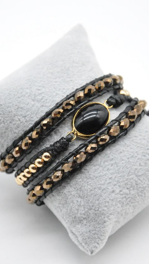 A stack of black leather bracelets with gold-toned chain links and a central black oval stone, displayed on a small gray fabric jewelry stand against a white background.