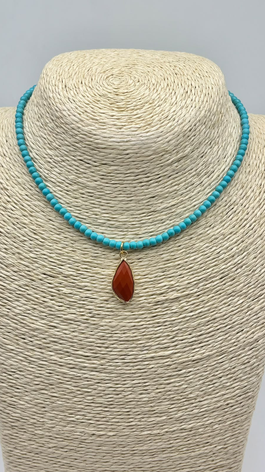 A turquoise beaded necklace with a teardrop-shaped reddish-brown stone pendant, displayed on a beige woven jewelry bust against a light gray background.