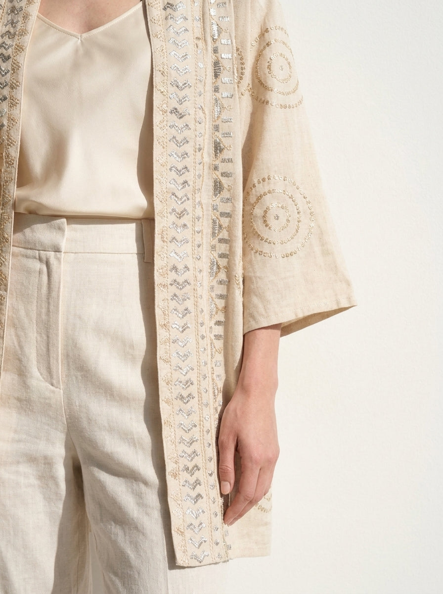 A close-up of a person wearing a cream-colored open-front cardigan with intricate silver embroidery and mirror work, paired with a matching cream satin top and linen trousers against a plain white background.