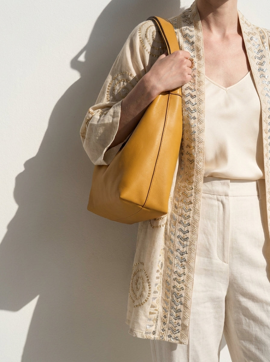 A person wearing a cream embroidered cardigan and white pants carries a large mustard yellow leather shoulder bag against a white wall with strong sunlight and shadows.
