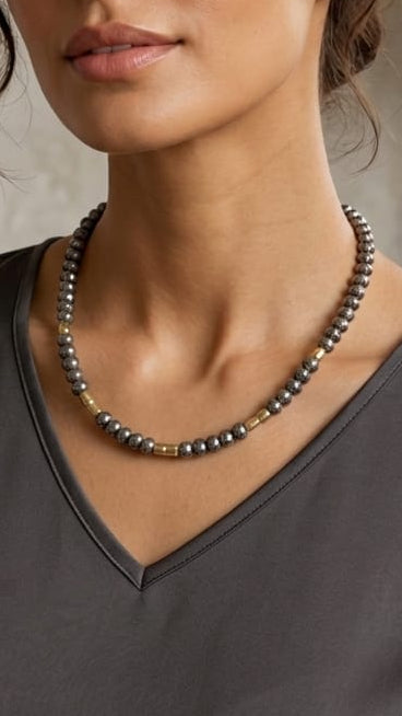 A woman wearing a dark gray satin V-neck top and a beaded pearl necklace, posing in a modern, neutral-toned interior setting.