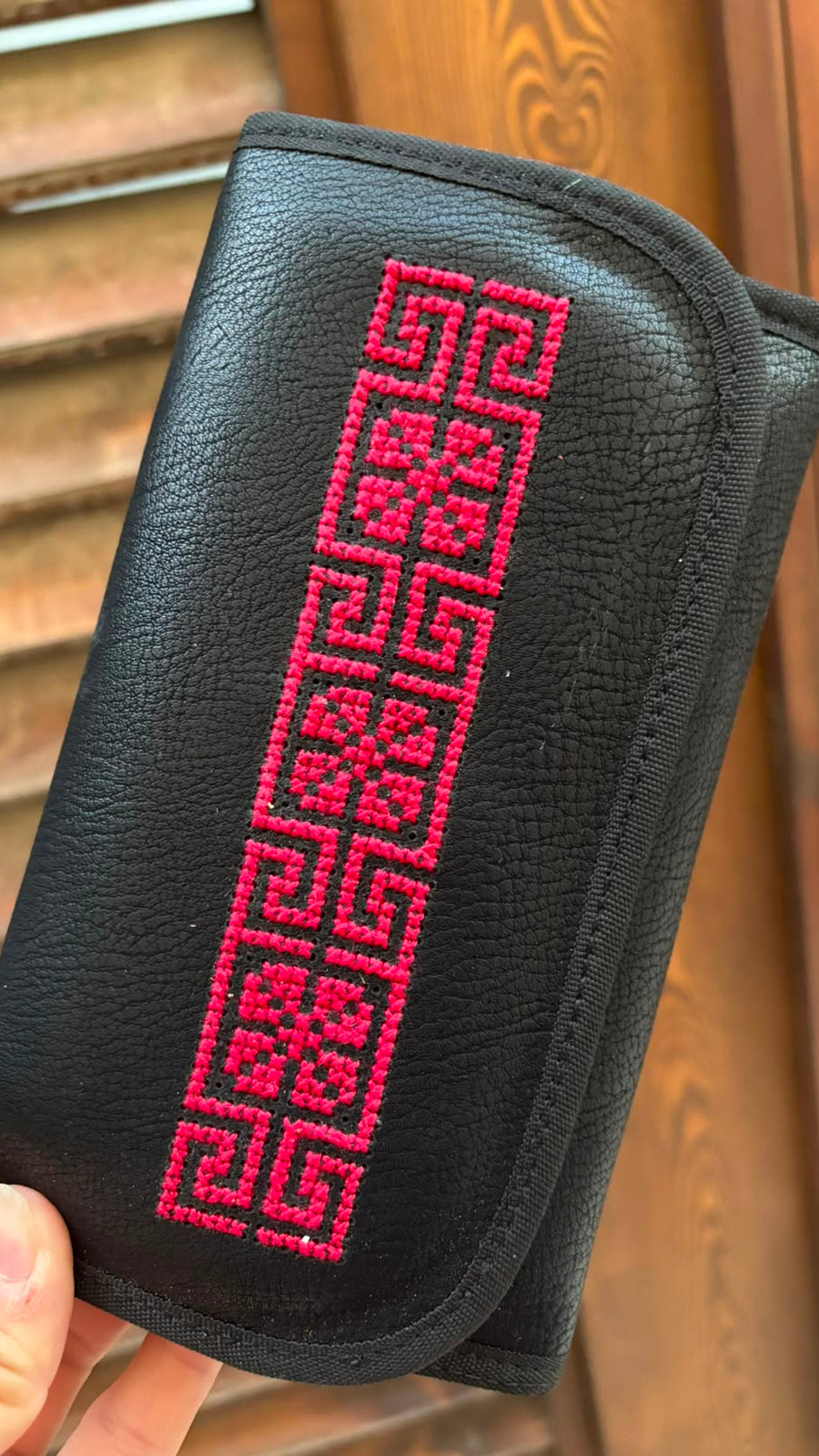 A black textured leather pouch with a bright pink geometric embroidered pattern, held by a hand in front of a wooden slatted door.