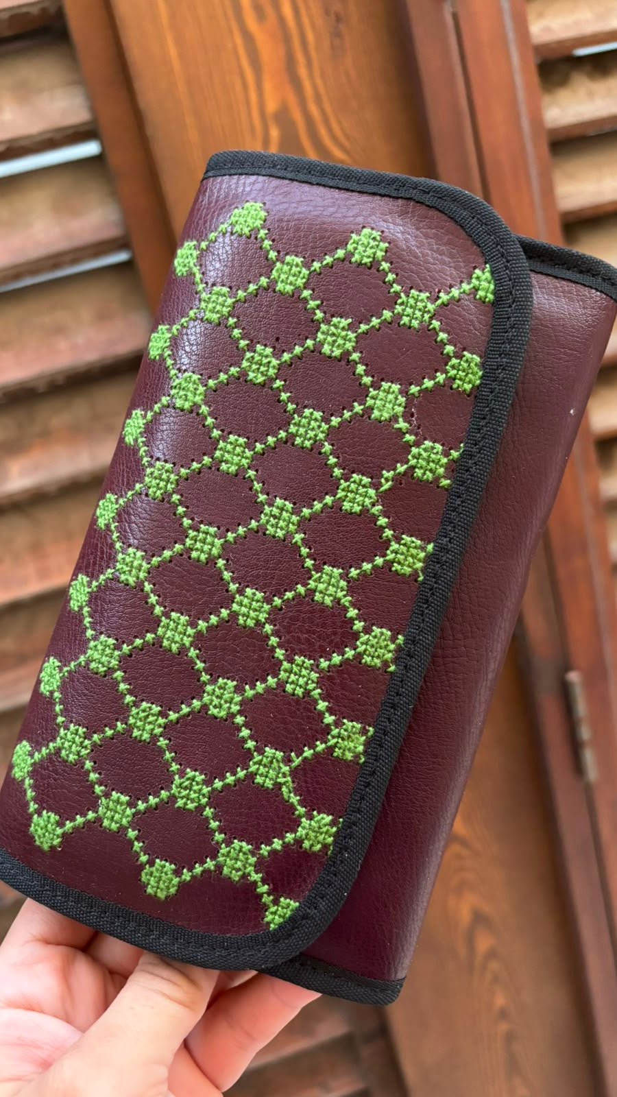 A hand holds a burgundy leather wallet or clutch with a green geometric patterned flap, set against a background of wooden shutters.