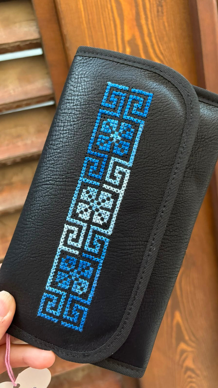 A black pebbled leather wallet or clutch with a vertical geometric blue and white embroidered pattern on the front flap, held by a hand in front of a wooden door.