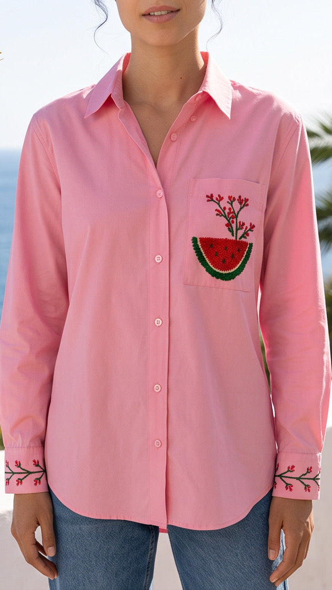 A woman wearing a pink long-sleeve button-up shirt with a watermelon patch embroidery on the pocket and floral embroidery on the cuffs, set against a sunny beach background with palm trees and the ocean.