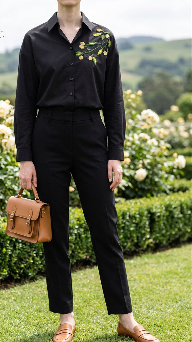 A model stands on a grassy lawn in front of a rose garden, wearing a black long-sleeve button-up shirt with green and yellow floral embroidery on the chest, paired with black trousers and tan loafers. She is holding a tan leather handbag.