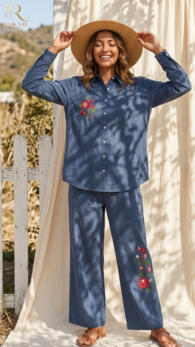 A woman modeling a matching blue long-sleeve button-up shirt and wide-leg pants set with red floral embroidery, styled with a straw hat and sandals in front of a cream fabric backdrop.