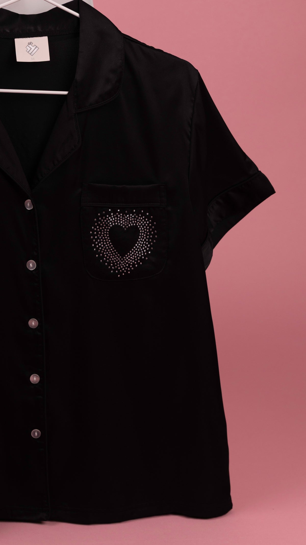 A black short-sleeve button-front shirt with a chest pocket featuring a heart-shaped rhinestone embellishment, hanging against a solid pink background.