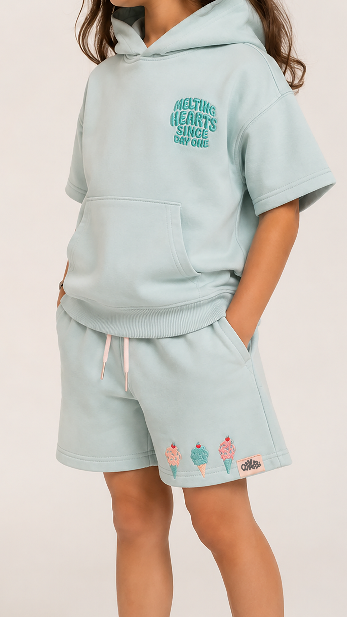 A child wearing a light mint green matching loungewear set, featuring a short-sleeve hoodie with a teal graphic on the chest and matching shorts with a pink drawstring and pastel ice cream cone embroidery.