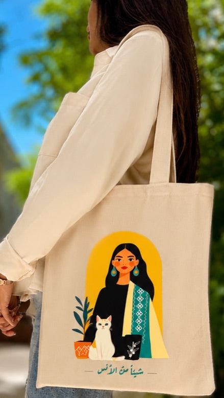 A woman in a white long-sleeve shirt carries a cream canvas tote bag outdoors. The bag features a colorful graphic illustration of a woman with a cat and Arabic text.