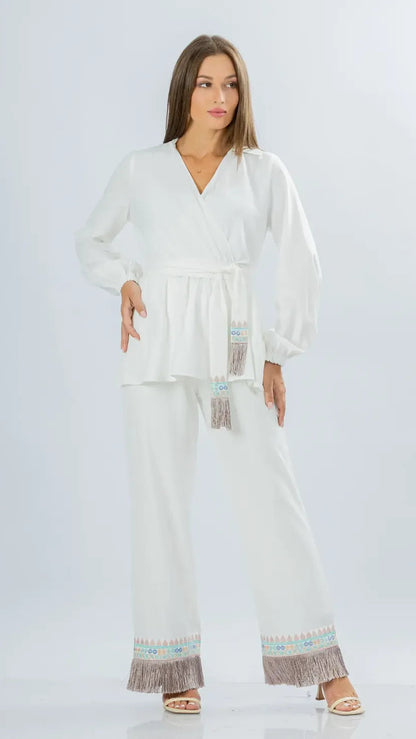 A woman modeling a white two-piece outfit consisting of a long-sleeve belted wrap top and matching wide-leg pants, both featuring light blue and brown patterned fringe trim at the hem.
