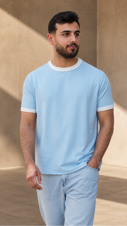 A man wearing a light blue ringer t-shirt with white trim on the collar and cuffs, paired with light blue jeans, standing in a modern architectural setting with warm beige walls and soft shadows.