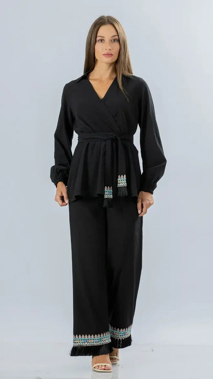 A woman modeling a black long-sleeve wrap-style top and matching wide-leg pants set with small multicolor embroidered details at the hem and waist, photographed against a light gray studio background.