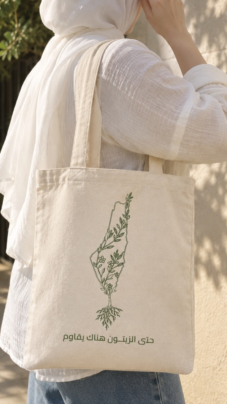 A person wearing a white hijab and white long-sleeve top carries a cream canvas tote bag with a green botanical illustration of the Palestinian flag and Arabic text reading 'Water is not the problem, the water carrier is.' The photo is taken outdoors against a light beige wall with soft shadows.