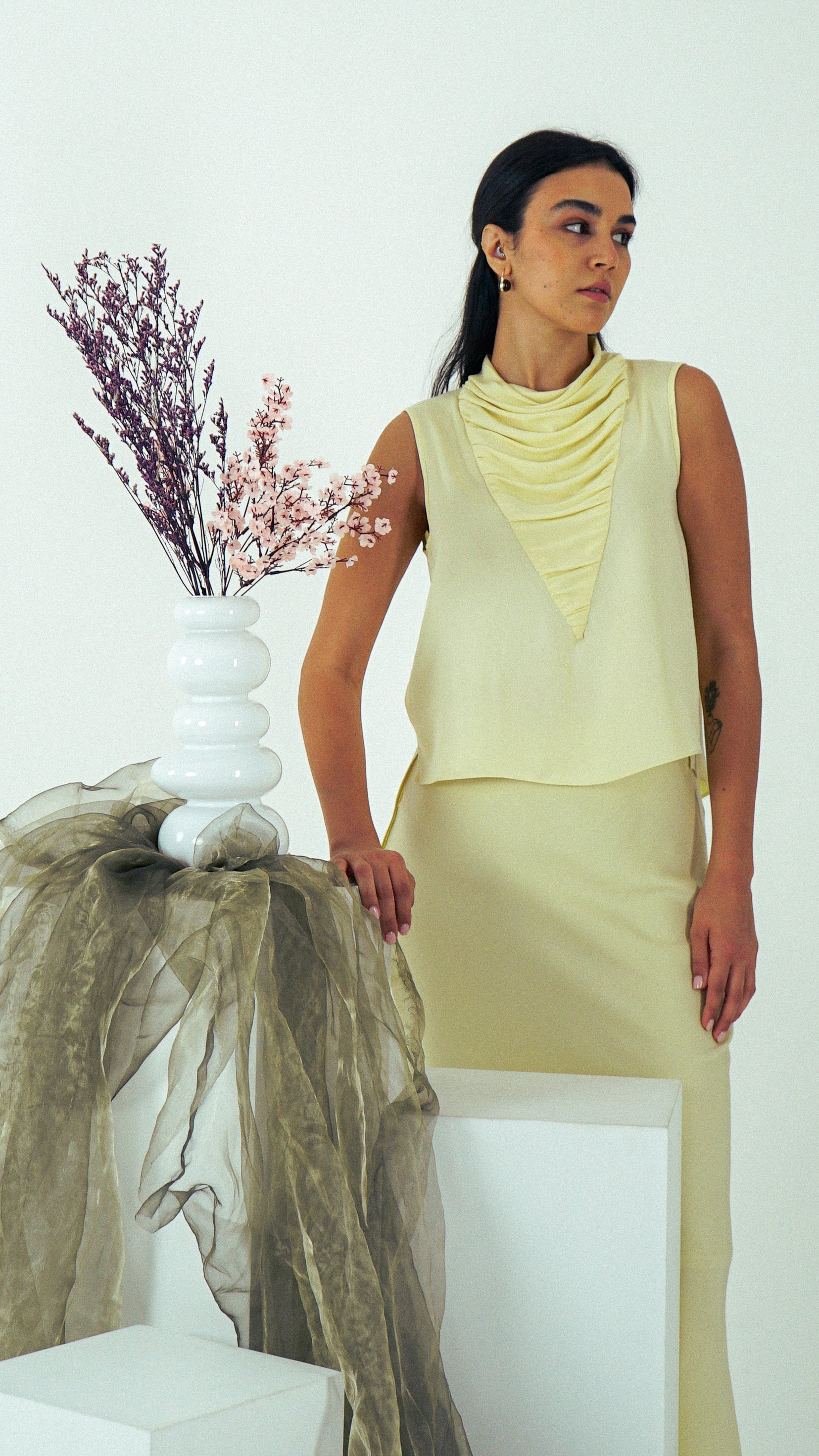 Woman wearing a light yellow sleeveless top and skirt standing next to a white vase with dried flowers on a white background