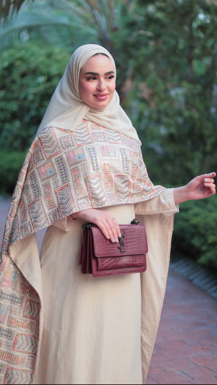 A woman wearing a beige hijab and a matching long-sleeve abaya with a geometric patterned shawl, holding a burgundy handbag, standing on a brick path surrounded by green foliage.
