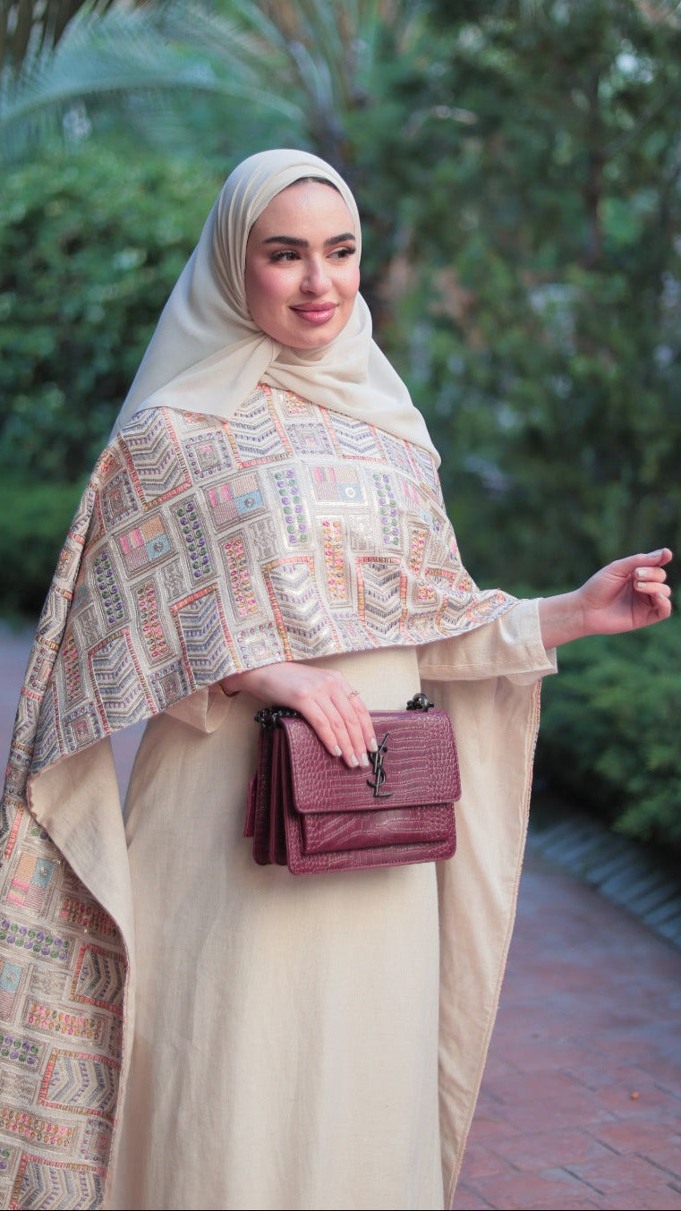 A woman wearing a beige hijab and a matching long-sleeve abaya with a geometric patterned shawl, holding a burgundy handbag, standing on a brick path surrounded by green foliage.