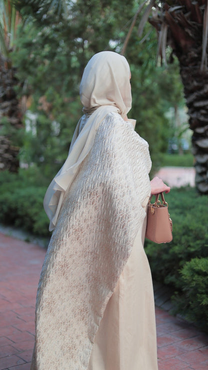 A woman wearing a cream-colored textured hijab and matching long dress, standing outdoors on a paved path with palm trees and greenery in the background.