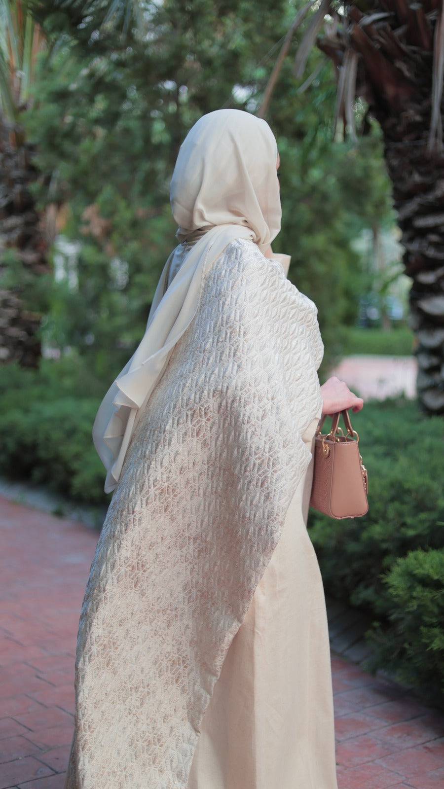 A woman wearing a cream-colored textured hijab and matching long dress, standing outdoors on a paved path with palm trees and greenery in the background.