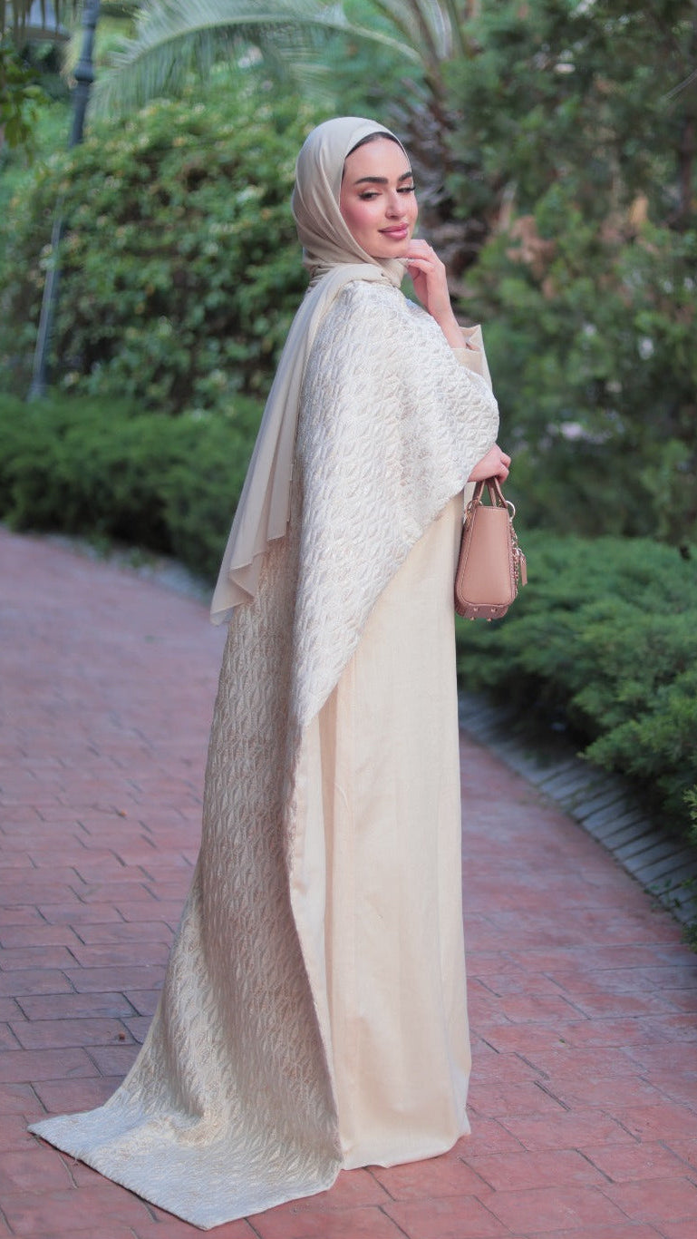 A woman wearing a cream-colored modest outfit with a matching hijab stands on a brick pathway surrounded by green bushes and palm trees, holding a small tan handbag.
