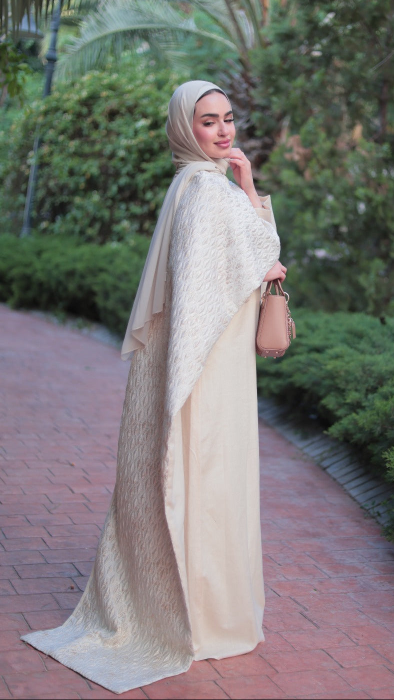 A woman wearing a cream-colored modest outfit with a matching hijab stands on a brick pathway surrounded by green bushes and palm trees, holding a small tan handbag.