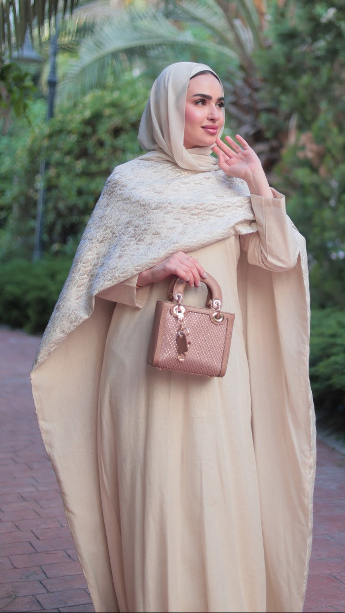 A woman wearing a beige abaya and matching hijab stands on a brick path in a garden, holding a small pink handbag.