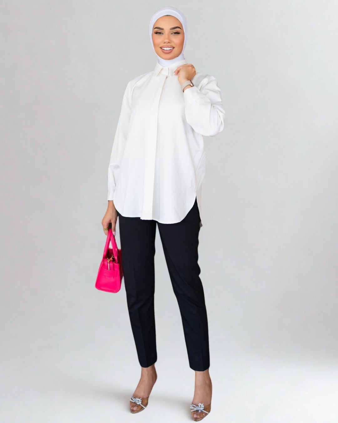 Woman wearing a white blouse, black pants, and a hijab, holding a pink handbag on a white background