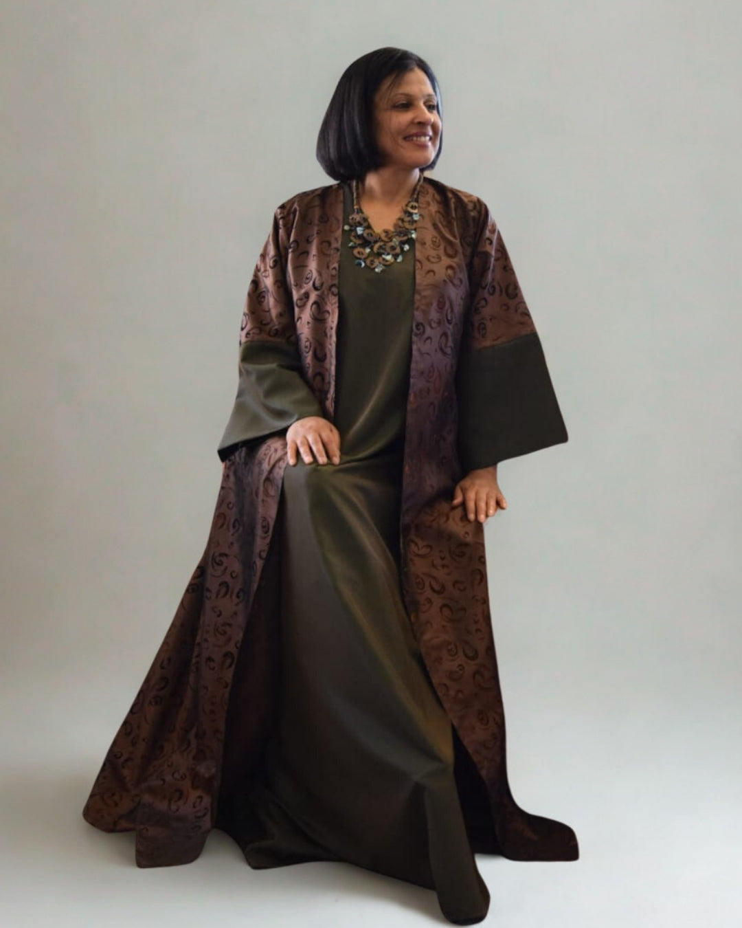 Woman wearing a long, dark green dress with a patterned brown shawl over it on a plain background