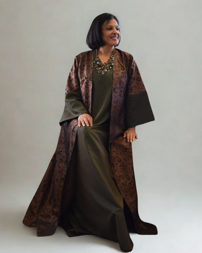 Woman wearing a long, dark green dress with a patterned brown shawl over it on a plain background