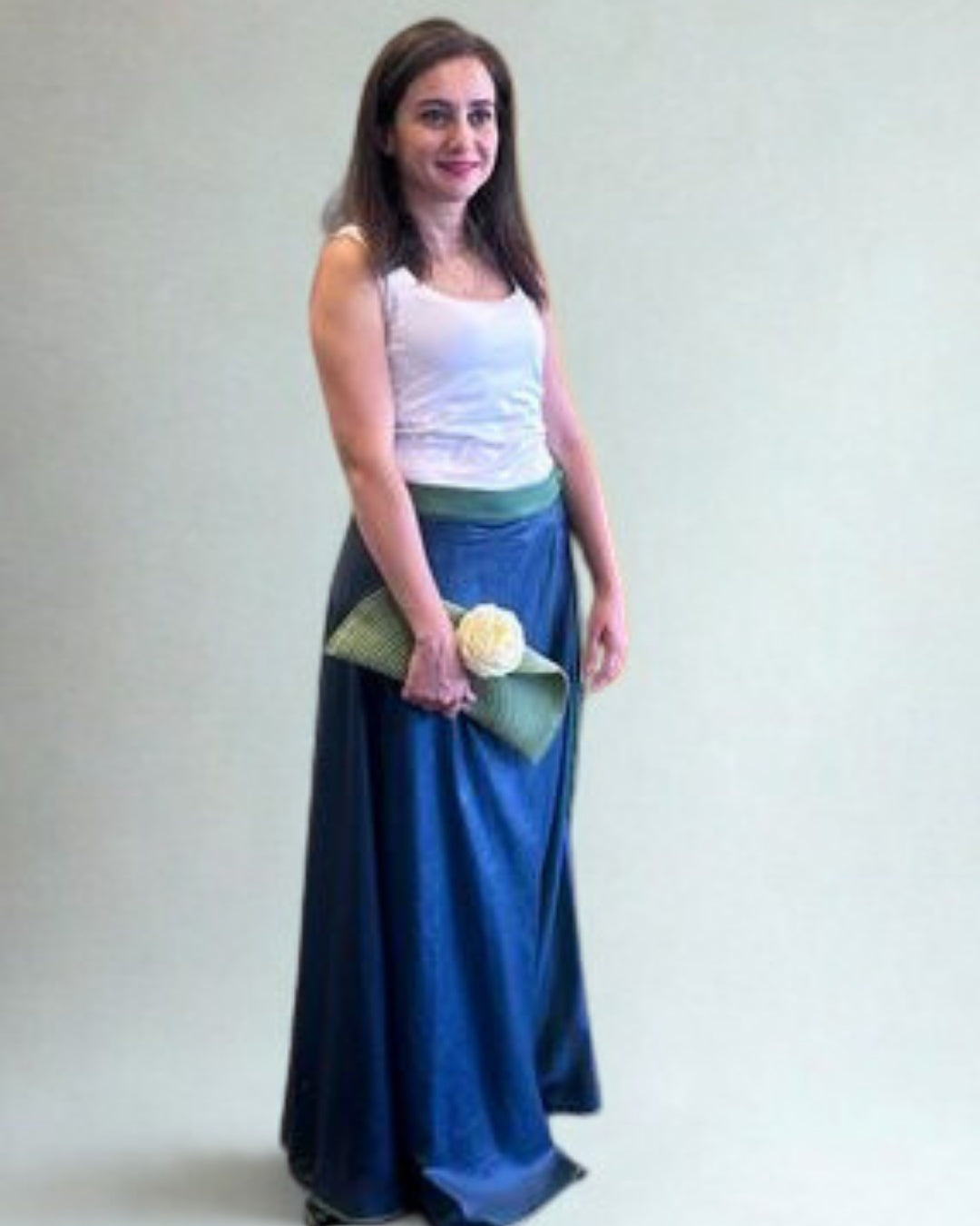 Woman wearing a white top and blue skirt holding a green clutch with a white flower against a plain background