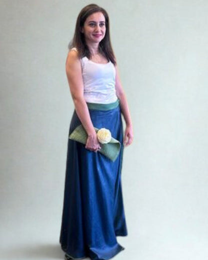Woman wearing a white top and blue skirt holding a green clutch with a white flower against a plain background