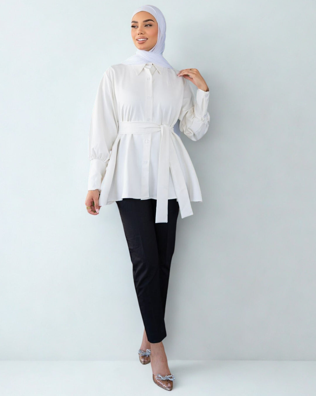 Woman wearing a white blouse and black pants with a hijab on a light gray background