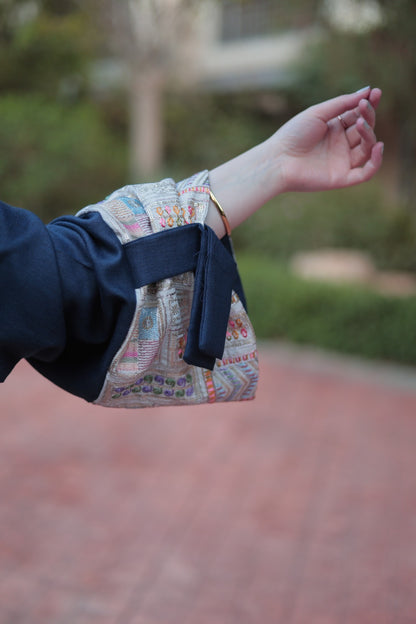 A close-up of a person's outstretched arm wearing a navy blue sleeve with a patterned fabric cuff and a gold bangle, set against a blurred outdoor background.