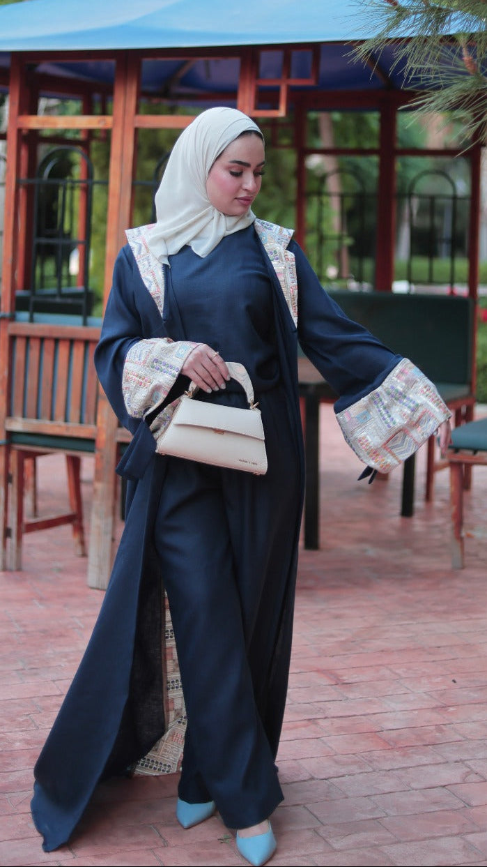 A woman wearing a dark navy blue modest outfit with beige patterned accents, a white hijab, and light blue shoes, standing in a paved outdoor area in front of a wooden gazebo with a blue roof.