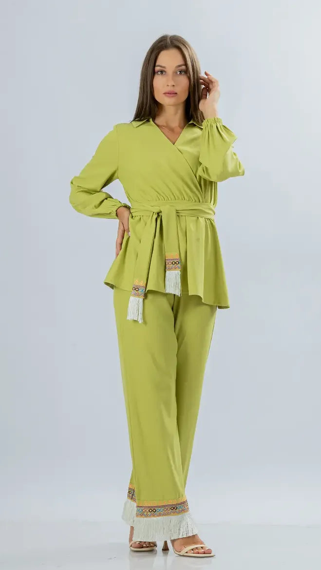 A woman modeling a bright lime green two-piece outfit consisting of a long-sleeve belted wrap-style top and matching wide-leg pants with decorative fringe and embroidered details near the hem, photographed against a light gray studio background.