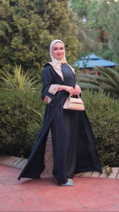 A woman wearing a black abaya and a beige hijab stands in a garden setting, holding a small beige handbag.