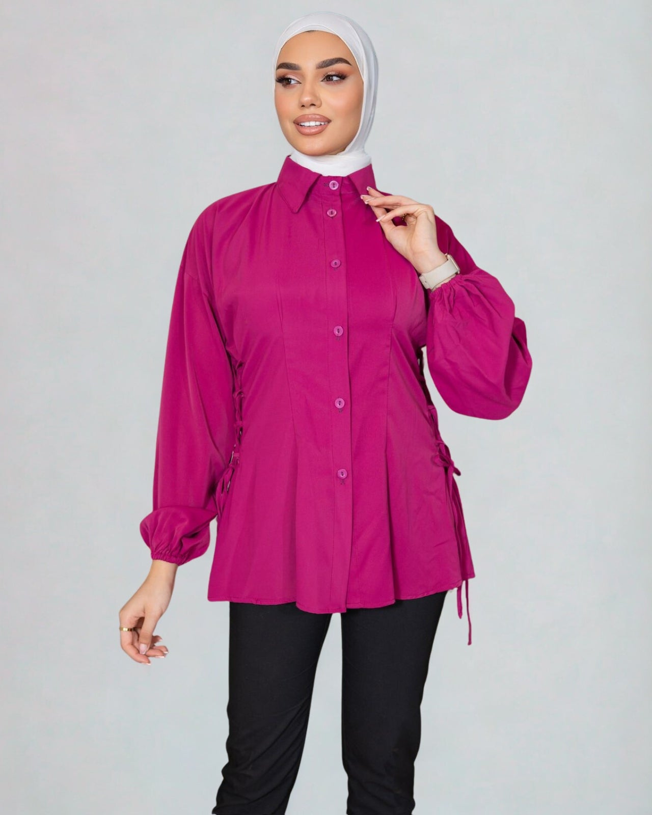 Woman wearing a bright pink blouse with long sleeves and a white hijab on a gray background