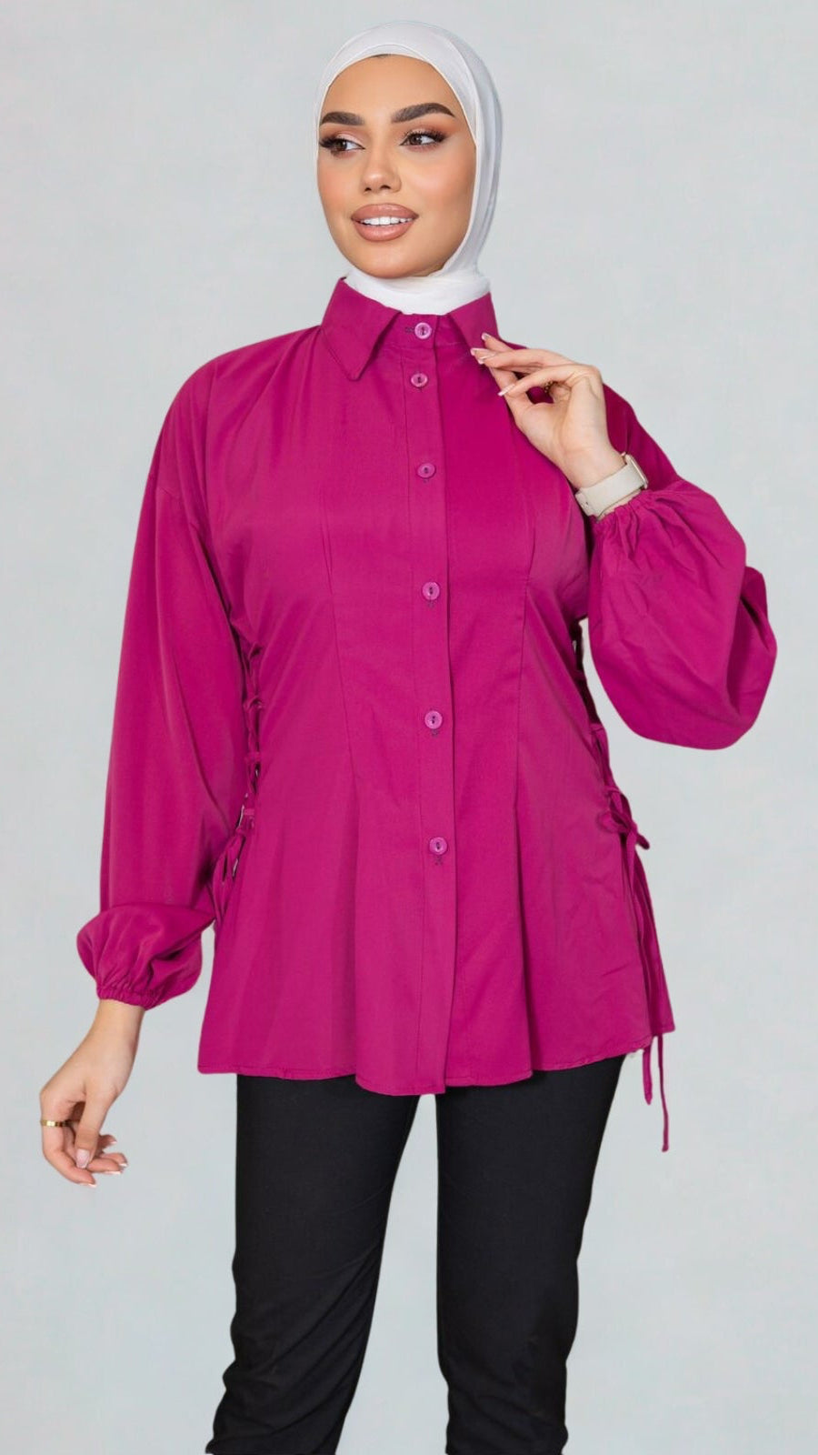 Woman wearing a bright pink blouse with long sleeves and a white hijab on a gray background