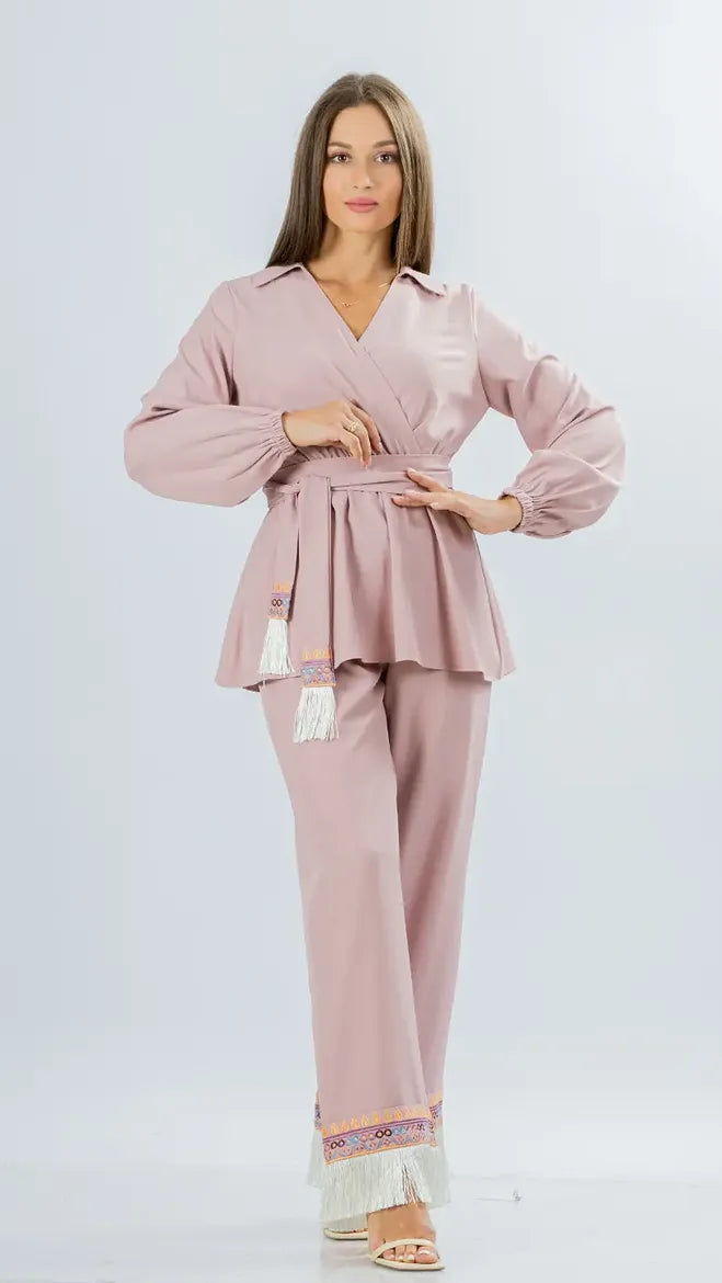 A woman modeling a dusty pink two-piece lounge set consisting of a long-sleeve belted wrap top and matching wide-leg pants with fringe and embroidered details at the ankles, set against a light gray studio background.