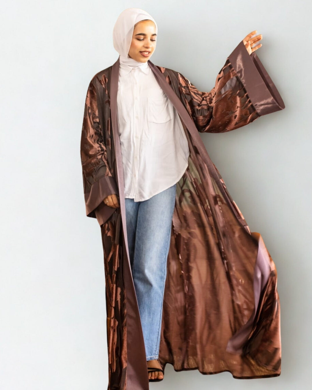 Person wearing a long brown coat over a white top and blue jeans on a light gray background