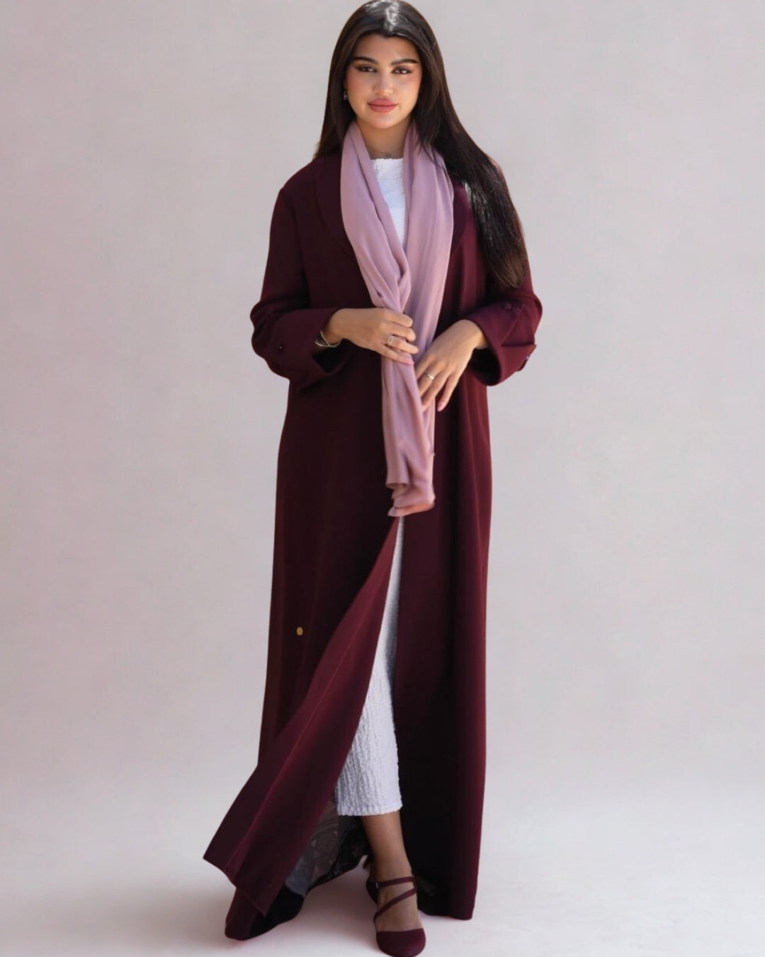 Woman wearing a long burgundy coat over a white outfit with a pink scarf on a plain background