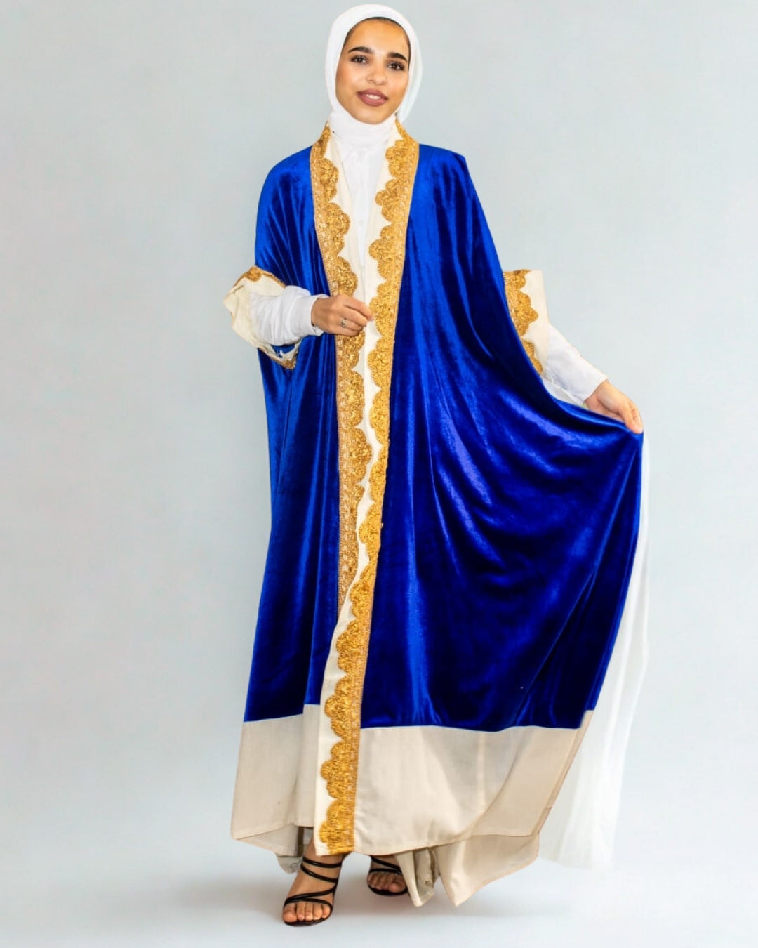 Woman wearing a blue and gold abaya with a white hijab on a gray background