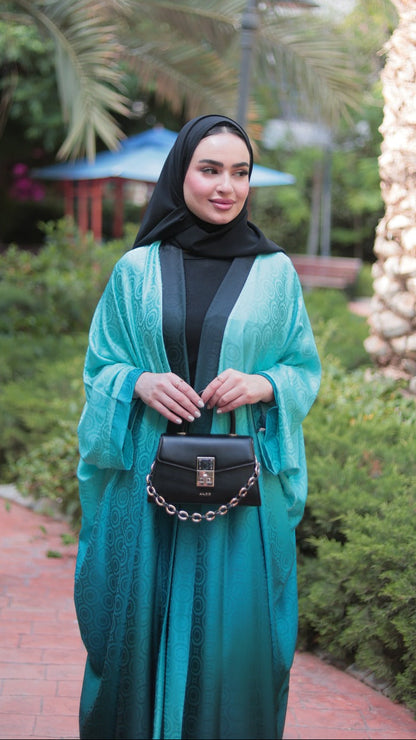A woman wearing a teal and green gradient abaya with a black inner layer and black hijab stands on a brick path in a garden with palm trees. She is holding a black crossbody bag with a gold clasp.
