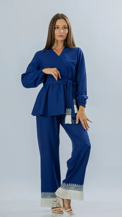 A woman modeling a royal blue two-piece set consisting of a long-sleeve belted wrap-style top and matching wide-leg pants, both finished with white fringe and blue patterned trim at the hem, photographed against a light gray studio background.