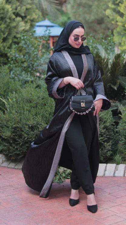 A woman wearing a black abaya with gray side panels, a black hijab, and black tights stands on a brick pathway in a garden, holding a small black handbag. A small dog is standing nearby on the right.