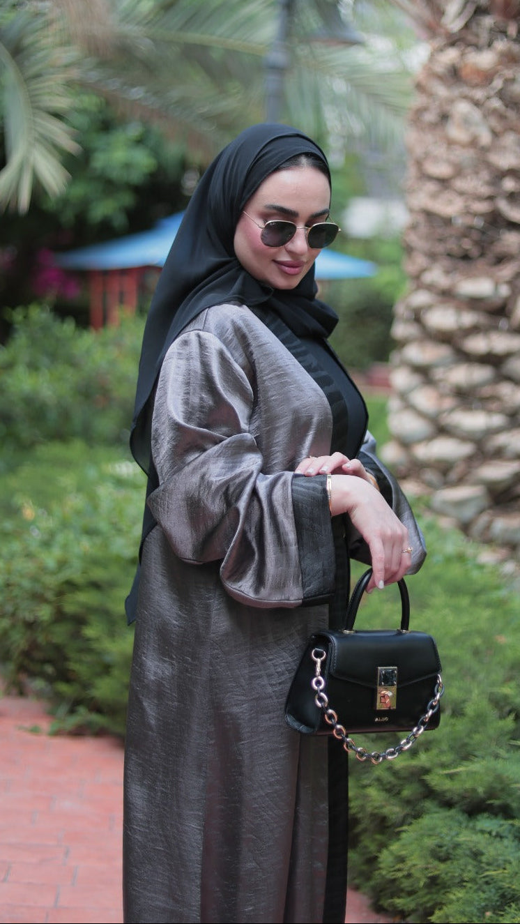 Woman in a gray abaya and black hijab holding a black handbag in a garden setting.