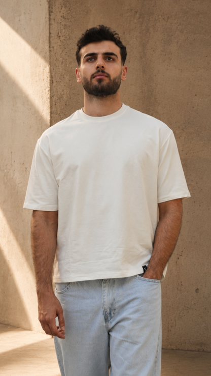 A man wearing a plain white crew-neck t-shirt and light blue jeans, standing against a textured concrete wall with diagonal sunlight shadows.